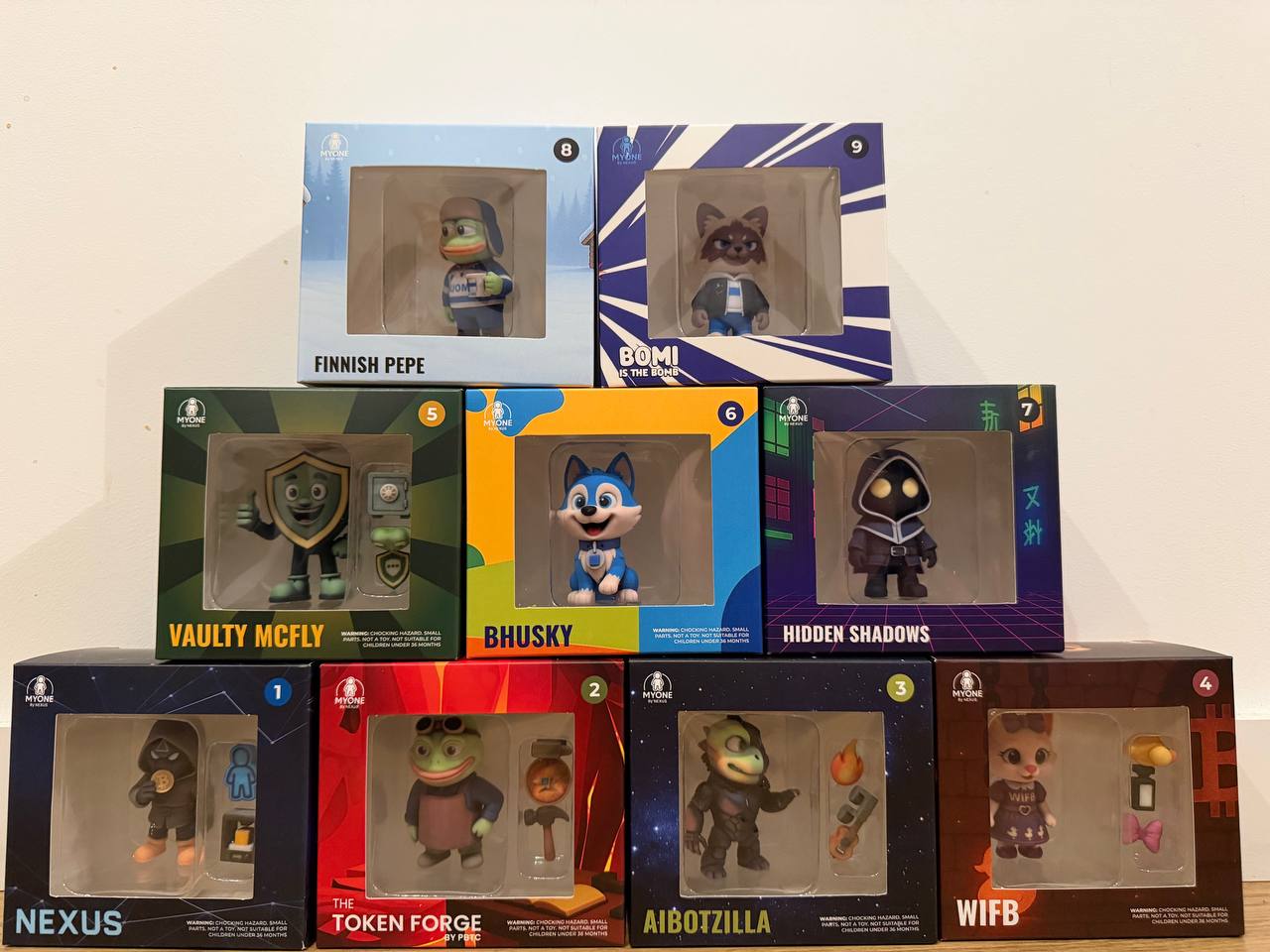 Photo of several boxed MYONE collectible figurines displayed together, including characters inspired by Token Forge, WIFB, AiBotZilla, and other crypto community projects